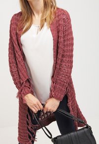 Un long cardigan en maille ouverte marron avec des franges. Porté sur un haut blanc, associé à un pantalon noir et un petit sac à main noir.