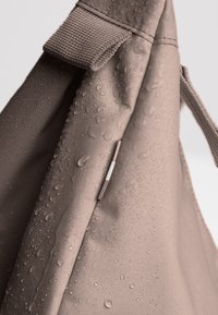 Borsa beige resistente all'acqua con una superficie testurizzata. Presenta gocce sul tessuto e un accento argento vicino alla parte superiore. Design compatto e pratico.