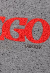 Grijze stof met een gespikkelde textuur, met grote, fuzzy rode letters die "GO" spellen en een bruin logo "J&Joy" eronder.