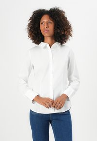 Camicia bianca a maniche lunghe con colletto classico, texture liscia e maniche con polsini, abbinata a jeans blu scuro.