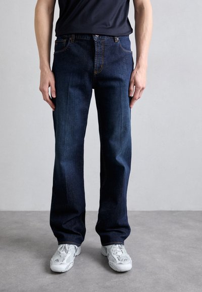 Jean en denim bleu foncé avec une coupe droite, dotés de coutures oranges et d'un style à cinq poches, associés à des chaussures de sport blanches.