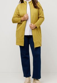 Chaqueta amarilla acolchada con un cuello alto, cierre de cremallera y bolsillos laterales, llevada sobre una blusa blanca y combinada con jeans azules y botas beige.