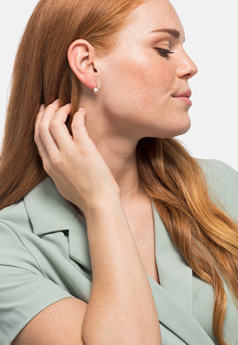 Orecchini a cerchio dorati con un piccolo accento di perla, indossati da una persona con lunghi capelli rossi, su una maglietta verde chiaro. Texture liscia e design elegante.