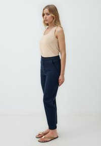 Top in beige, pantaloni navy, taglio slim fit, tessuto morbido, sandali piatti con dettagli dorati, in posa in piedi su uno sfondo bianco semplice.