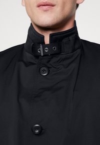 Schwarze wasserdichte Jacke mit hohem Kragen, zwei großen Knöpfen und verstellbaren Beschlägen. Glattes Material mit einem schlanken Design.