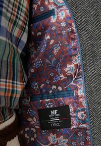 Texturiertes Fischgrat-Jacket mit einem bordeauxroten, floralem Innenfutter, das blaue Akzente und ein Textilschild mit der Aufschrift "NOVA FIDES Italian Fabric" aufweist.