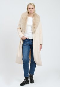 Lang crèmekleurige jas met een nepbont kraag, gedragen over een witte top en blauwe jeans. Het model staat met één hand in de zak en draagt zwarte enkellaarsjes.