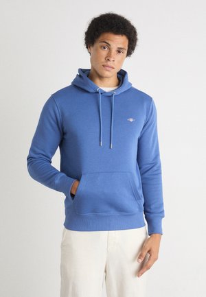 Junger Mann trägt einen blauen Hoodie mit Fronttasche und cremefarbene Hose, steht vor einem einfarbigen hellen Hintergrund.