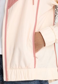 En persons hand inne i sidofickan på en ljusrosa jacka med elastiska ärmslut och mörkblå jeans synliga längre ner.