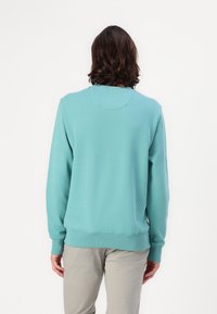 Tirkizna dukserica s okruglim izrezom, rebrastim manžetama i rubom. Mekani materijal, jednostavan dizajn, prikazana s leđa. Nosena sa svijetlosivim hlačama.