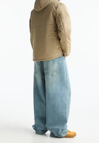 Beige gewatteerd jack met capuchon en een losse pasvorm, gecombineerd met lichtblauwe wijde spijkerbroeken en gele sneakers, gezien vanaf de achterkant.