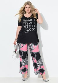 T-shirt nera con scollo a V e testo grafico bianco; abbinata a pantaloni larghi rosa e neri con un motivo a fantasia, che presentano stampa animale e dettagli a catena.