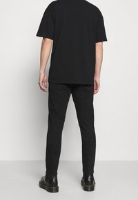 Person stående vänd bort från kameran, iklädd en svart kortärmad skjorta, svarta byxor och svarta läderkängor mot en vanlig bakgrund.