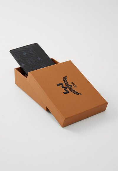 En sort teksturert lommebok delvis synlig i en brun pappeske, med MCM-logoen i svart, i en slank rektangulær form.