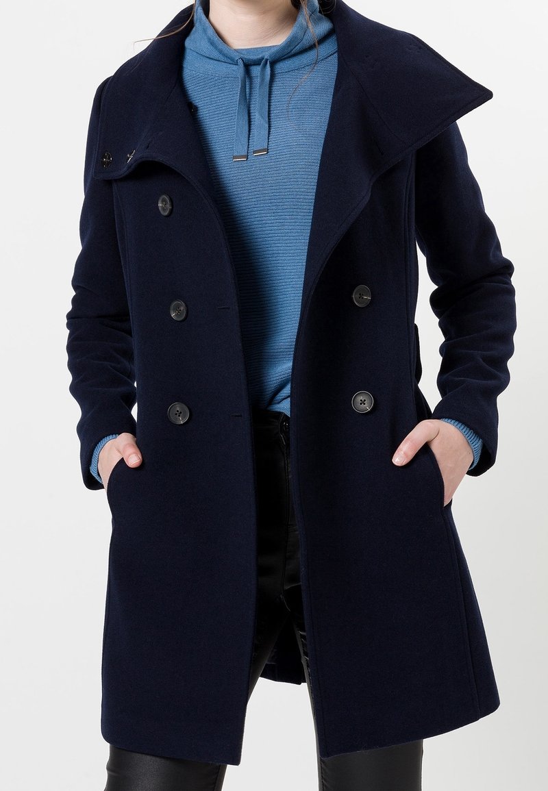 Femme portant un manteau bleu marine croisé par-dessus un sweat à capuche bleu, les mains dans les poches du manteau.