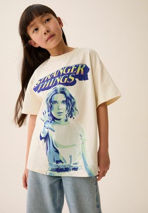 Ljusbeige oversized t-shirt med en blå grafisk tryck av en karaktär från "Stranger Things" med en hand upplyft, tillsammans med blå jeans.