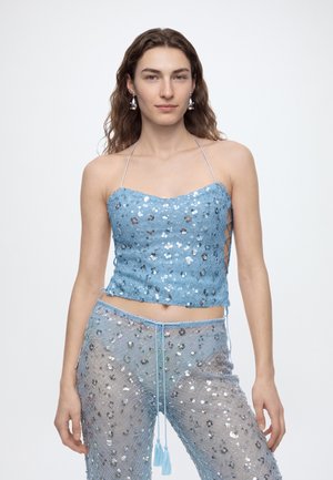 Personne portant un crop top halter bleu à sequins et un pantalon semi-transparent assorti avec des accents argentés, debout devant un fond blanc.