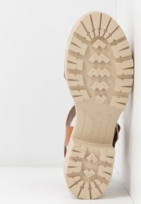 Semelle extérieure en caoutchouc beige d'une sandale marron clair avec des sculptures profondes et des rainures décoratives, présentée sur un fond blanc.