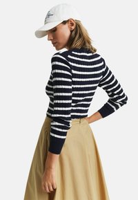 Streifenpullover in Marineblau und Weiß mit Rippenstruktur, langärmlig, kombiniert mit einem beigen, hoch taillierten Rock. Das Modell trägt eine weiße Mütze.