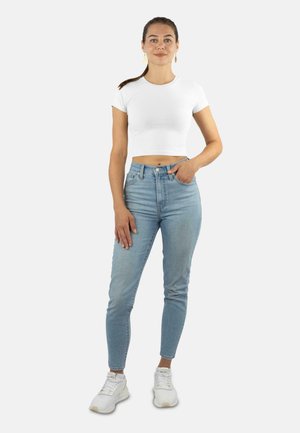 Weißes, kurzes T-Shirt, hoch sitzende hellblaue Jeans und weiße Sneakers. Das Outfit zeichnet sich durch ein figurbetontes Design mit einer glatten Textur aus.