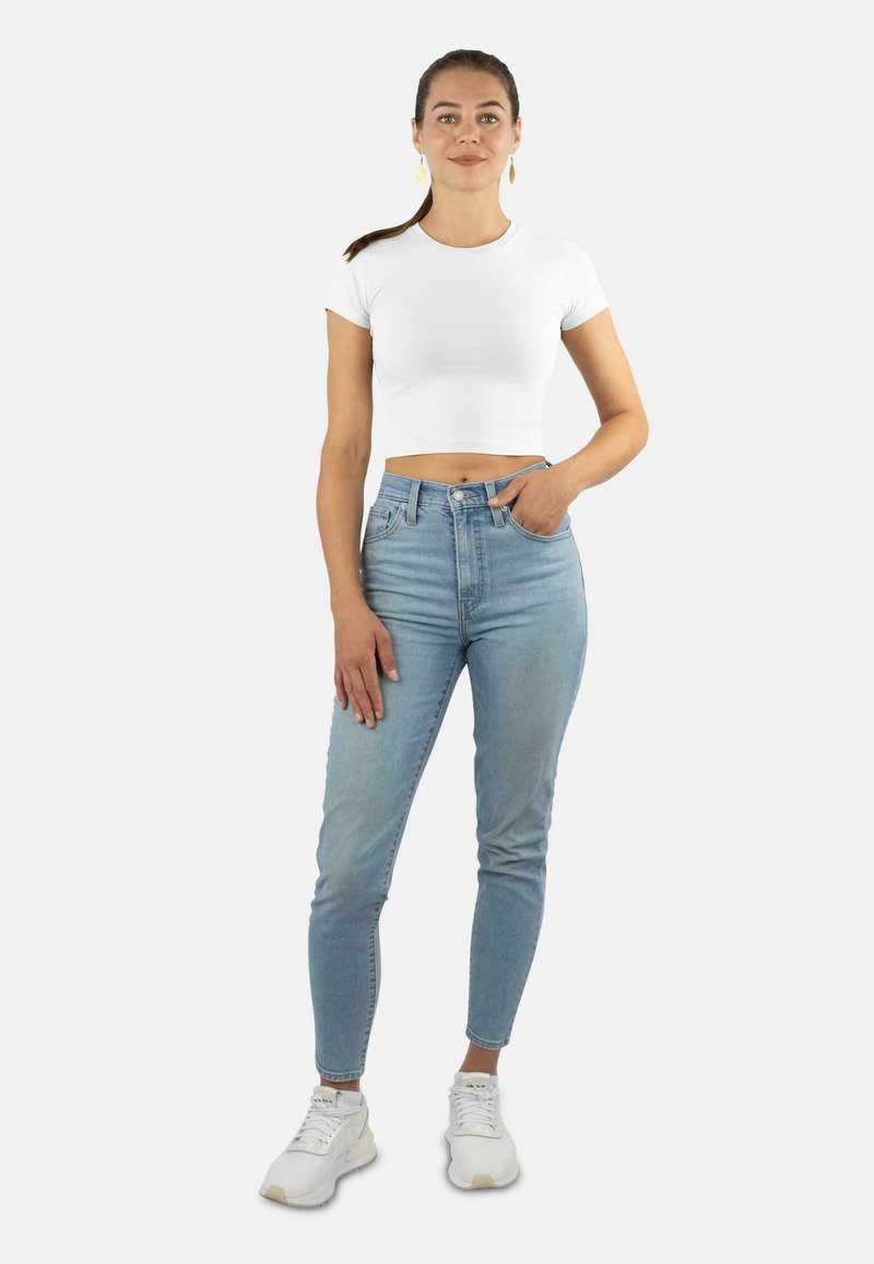 Weißes, kurzes T-Shirt, hoch sitzende hellblaue Jeans und weiße Sneakers. Das Outfit zeichnet sich durch ein figurbetontes Design mit einer glatten Textur aus.