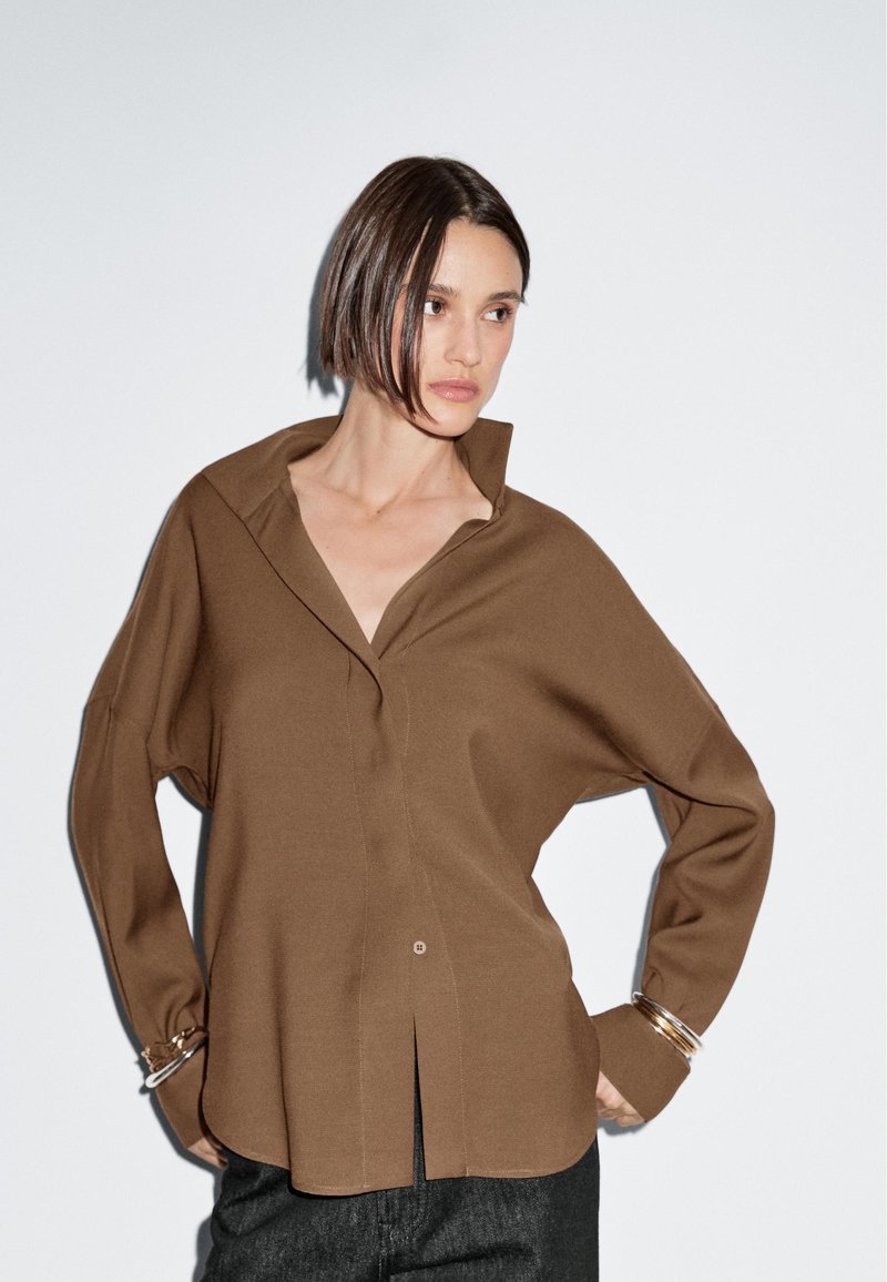 Brown collared shirt with a relaxed fit, featuring a button front and side split. Made from soft fabric; has long sleeves and minimal detailing.