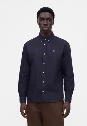 Homme portant une chemise boutonnée bleu marine foncé avec un petit logo vert sur la poitrine et un pantalon marron, debout devant un fond blanc uni.