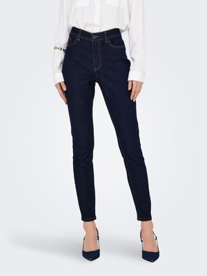 Kvinde iført mørkeblå slim fit jeans, hvid bluse med lange ærmer og mørke spidse hæle, stående mod en ensfarvet baggrund.