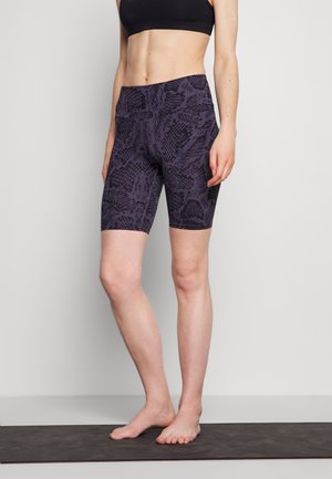 Frau steht barfuß auf einer Yogamatte und trägt einen schwarzen Sport-BH sowie gemusterte dunkelgraue, hochtaillierte Radlerhosen.