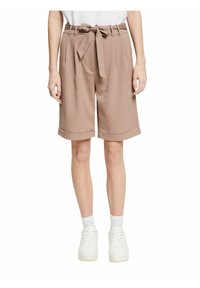 Bermudas beige a medida con un cinturón atado, pliegues y dobladillos. Llevadas con zapatillas blancas y calcetines para un look casual.