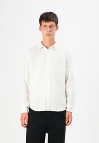 Camisa blanca texturizada con botones en la parte frontal, cuello clásico y mangas largas, combinada con pantalones negros.