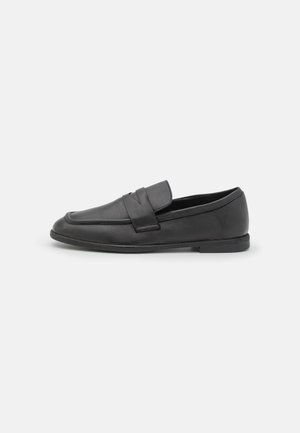 Mocassins en cuir noir présentant un design cousu, un talon bas et une bride sur le dessus pour le détail. Texture lisse, forme classique.