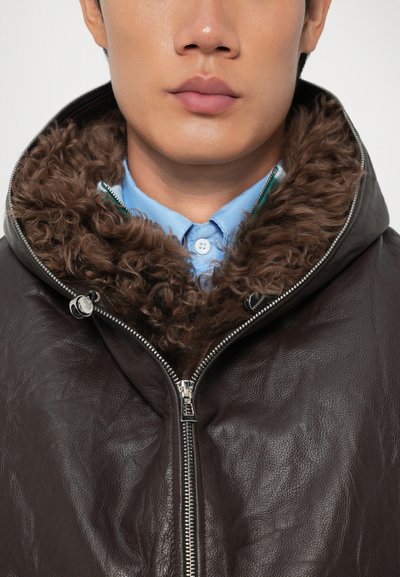 Man wearing dark brown leather jacket with large curly fur collar over light blue button-up shirt, close-up on chest and lower face.