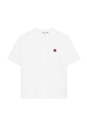 Uomo che indossa una camicia bianca a maniche corte con logo rosso, foulard fantasia al collo, pantaloni blu scuro e cappello blu scuro con scritta bianca su sfondo uniforme.