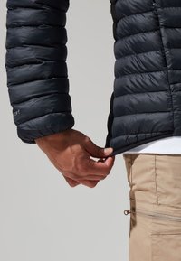 Jūras spēku pūkains mētelis ar quilted tekstūru un elastīgām piedurknēm, kas tiek pielāgots apakšmalai. Redzamā rāvējslēdzēja kabata uz gaiši brūnām biksēm.