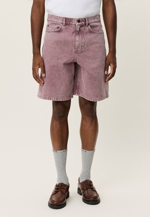 Verblasste pinke Jeansshorts mit Seitentaschen, Gürtelschlaufen und geradem Bein. Kombiniert mit grauen Socken und braunen Schuhen.