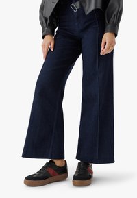 Person in dunklen, weiten Jeans, schwarzen Sneakers mit roten Streifen und einer schwarzen Lederjacke, steht vor einem weißen Hintergrund.