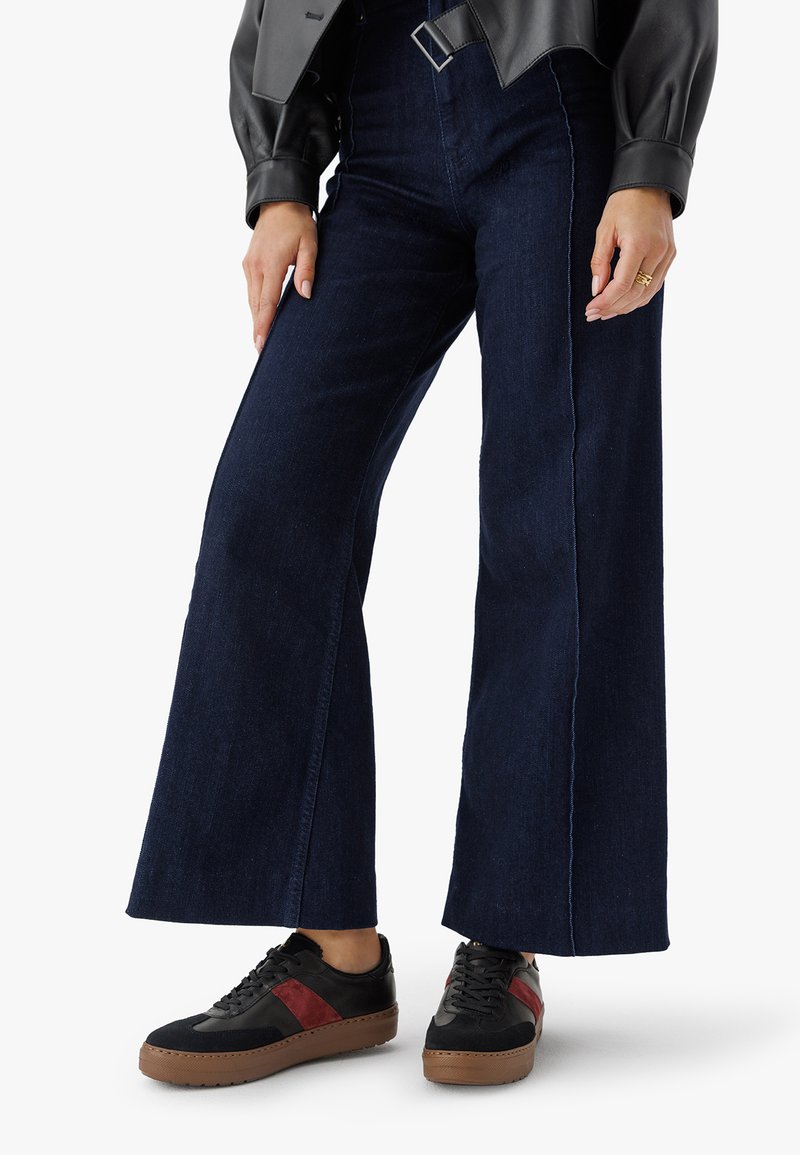 Person in dunklen, weiten Jeans, schwarzen Sneakers mit roten Streifen und einer schwarzen Lederjacke, steht vor einem weißen Hintergrund.