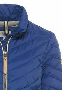 Sininen tikkikangas takki, jossa on korkea kaulus, vetoketjullinen etuosa ja beigeitä yksityiskohtia. Ominaisuuksiin kuuluvat vinot tikkaus ja ripustuslenkki kauluksessa.