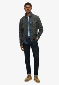 Grå zip-up jakke med forlommer, lagdelt over en denimskjorte og hvid t-shirt, parret med mørke jeans og lysebrune ruskindssko.