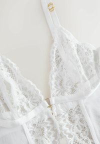 Lencería blanca de encaje con delicados patrones florales, malla transparente, diseño de escote en V y un detalle de herrajes en tono dorado en el centro.