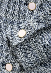 Tessuto blu e bianco strutturato con un motivo a maglia, caratterizzato da grandi bottoni bianchi e dorati sull'orlo e nell'area del colletto.