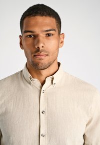 Jonge man met kort krullend haar en een getrimde baard, die recht vooruit kijkt tegen een effen achtergrond.