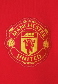 Un tessuto rosso presenta un ricamo in oro dello stemma del Manchester United. Il design include un leone e una nave, incorniciati da un bordo circolare.