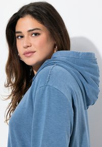 Blauer Kapuzenpullover mit lockerer Passform. Weicher Stoff, sichtbare Nähdetails und ein leicht ausgefranster Rand an der Schulter.