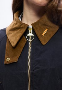 Κοντινό πλάνο ενός ατόμου που φοράει μ πλε σκούρο σακάκι με καφέ κολάρο από κορδόνι, χρυσό φερμουάρ και μικρή ορθογώνια καρφίτσα στο δεξί κολάρο που γράφει "BEDALE".