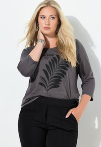 Pitkähiuksinen vaaleatukkainen nainen, joka pukeutuu harmaaseen yläosaan, jossa on musta lehtikuvio, ja mustiin housuihin, poseeraa käsi toisessa kaulalla ja toinen taskussa.