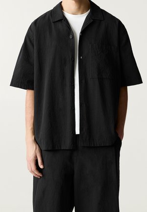 Mann trägt locker sitzendes schwarzes kurzärmeliges Hemd über weißem T-Shirt, kombiniert mit passenden schwarzen Hosen, steht vor schlichtem Hintergrund.