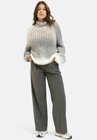 Frau, die einen grauen und weißen grob gestrickten Rollkragenpullover, graue weite Hosen und weiße Sneakers trägt, steht mit den Händen in den Taschen.