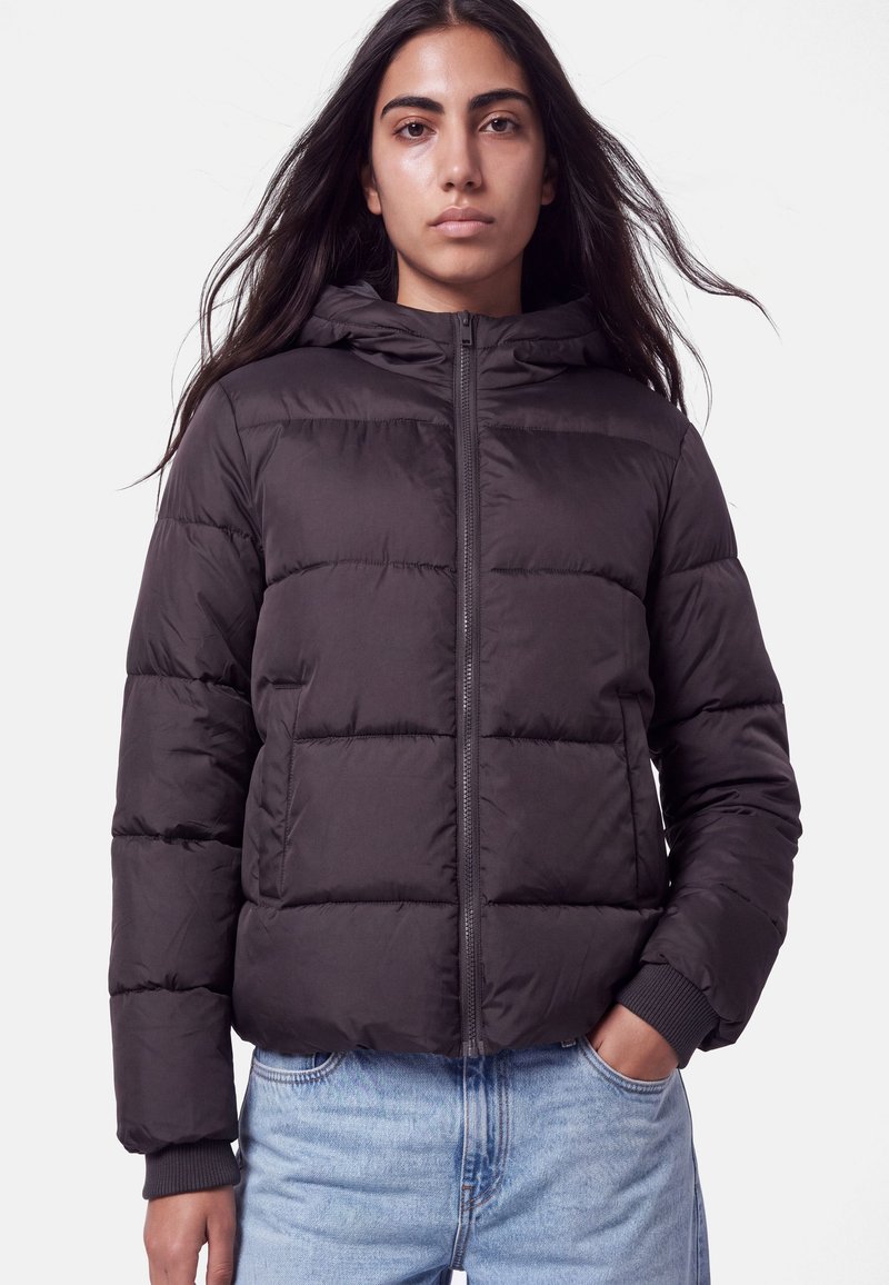 Kvinde med langt mørkt hår iført en sort dunjakke og lyseblå jeans, stående med den ene hånd i lommen mod en ensfarvet baggrund.